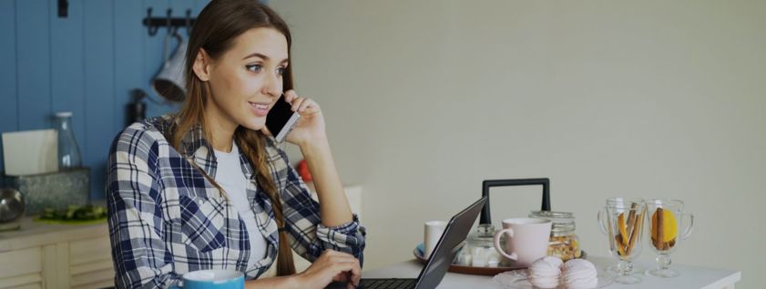 Young woman multitasking on phone and laptop in a cozy kitchen setting.
